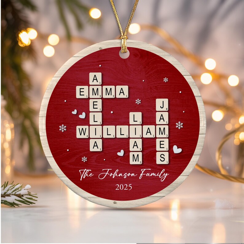 A round, red, wood-textured Christmas ornament featuring a crossword-style arrangement of letter tiles spelling AMELIA, EMMA, WILLIAM, and JAMES.