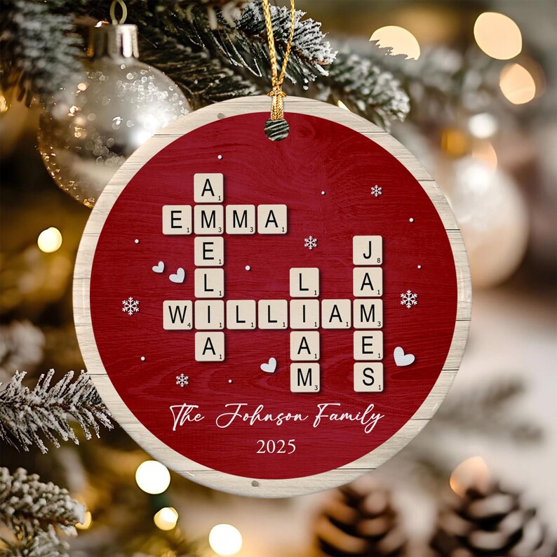 A round ceramic Christmas ornament with a red wood-grain background featuring a crossword-style arrangement of letter tiles spelling the names Emma, Amelia, William, Liam, and James. The text reads The Johnson Family 2025.