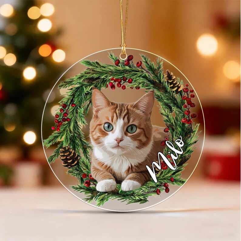 A round, clear acrylic ornament featuring a printed image of an orange and white tabby cat centered within a festive wreath of green pine needles, red berries, and brown pinecones. The name Milo is written in white script.