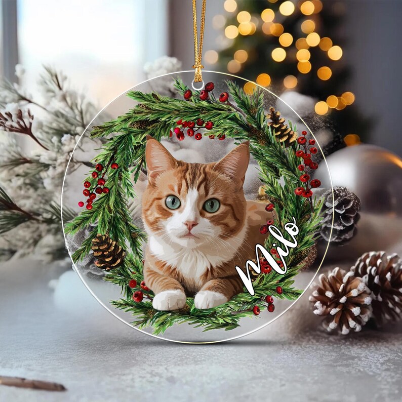 A circular Christmas ornament featuring a ginger and white tabby cat centered within a wreath of green pine needles, red berries, and small pinecones. The name Milo is written in white cursive script across the lower right side.