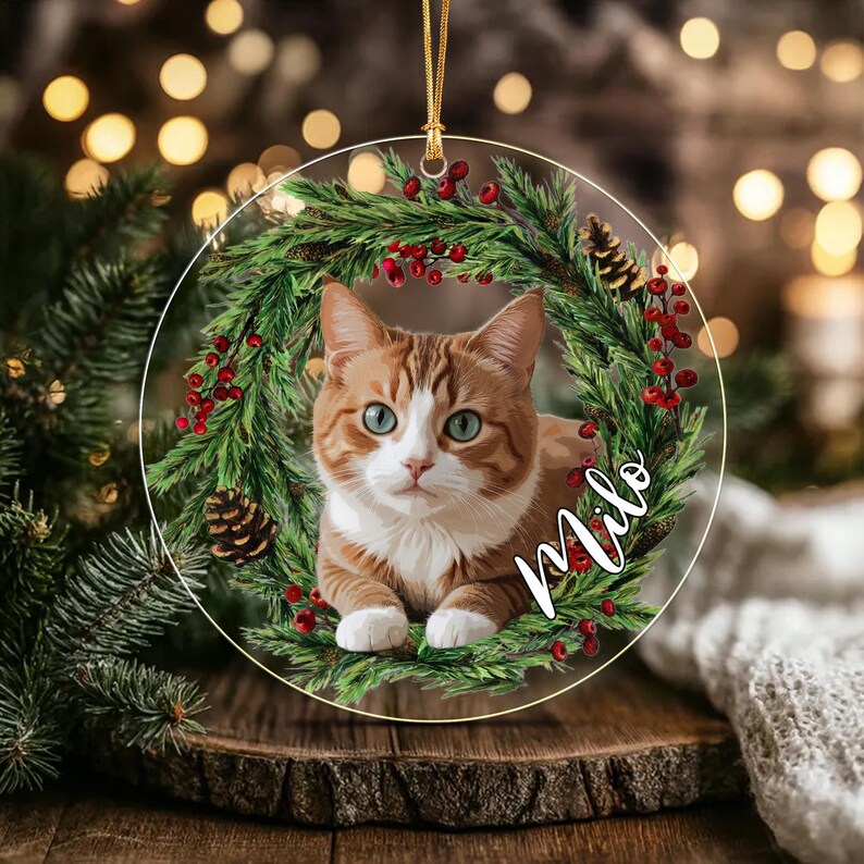 A round, clear acrylic Christmas ornament featuring a printed illustration of an orange and white tabby cat centered within a festive wreath of green pine needles, red berries, and pinecones.