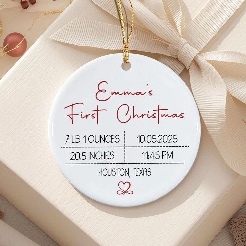 A round white ceramic ornament with a gold hanging string. The text reads Emma's First Christmas, 7 LB 1 OUNCES, 10.05.2025, 20.5 INCHES, 11:45 PM, and HOUSTON, TEXAS, with a small red heart symbol at the bottom.