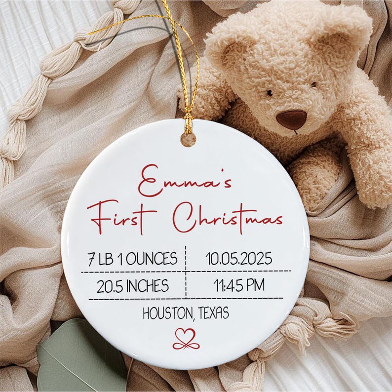 A round white ceramic ornament with a gold hanging string. The text reads Emma's First Christmas, 7 LB 1 OUNCES, 10.05.2025, 20.5 INCHES, 11:45 PM, and HOUSTON, TEXAS, with a red heart icon at the bottom center of the ornament.