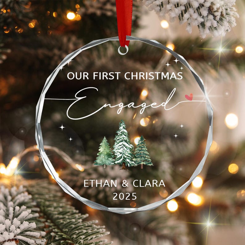 A circular clear glass ornament with faceted edges, featuring the text "OUR FIRST CHRISTMAS Engaged" with a small red heart, a watercolor illustration of three pine trees, and the names "ETHAN & CLARA" with the year "2025" below.