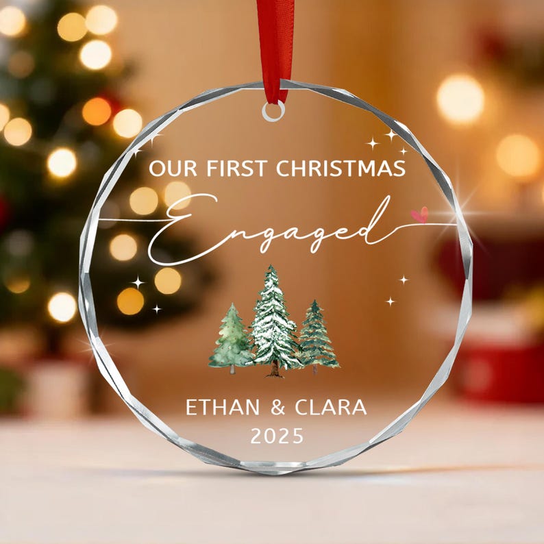 A circular clear glass Christmas ornament with faceted edges, featuring a red ribbon hanger. The text reads OUR FIRST CHRISTMAS Engaged, ETHAN & CLARA, and 2025.