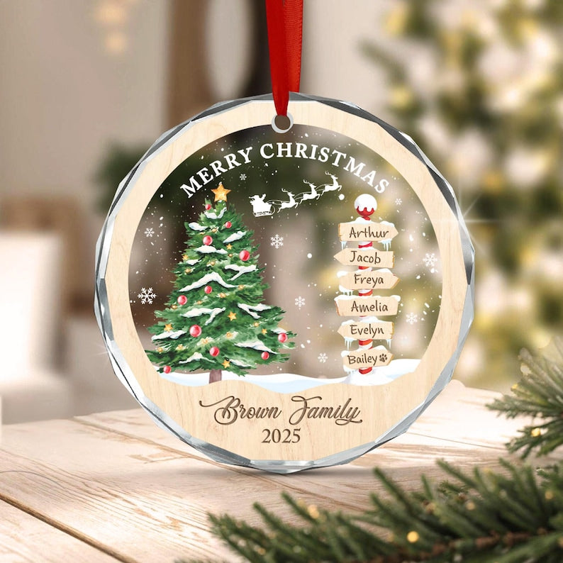 A round, faceted glass Christmas ornament with a red ribbon hanger. The design features a green Christmas tree, a silhouette of Santa and reindeer, and a signpost with the names Arthur, Jacob, Freya, Amelia, Evelyn, and Bailey.