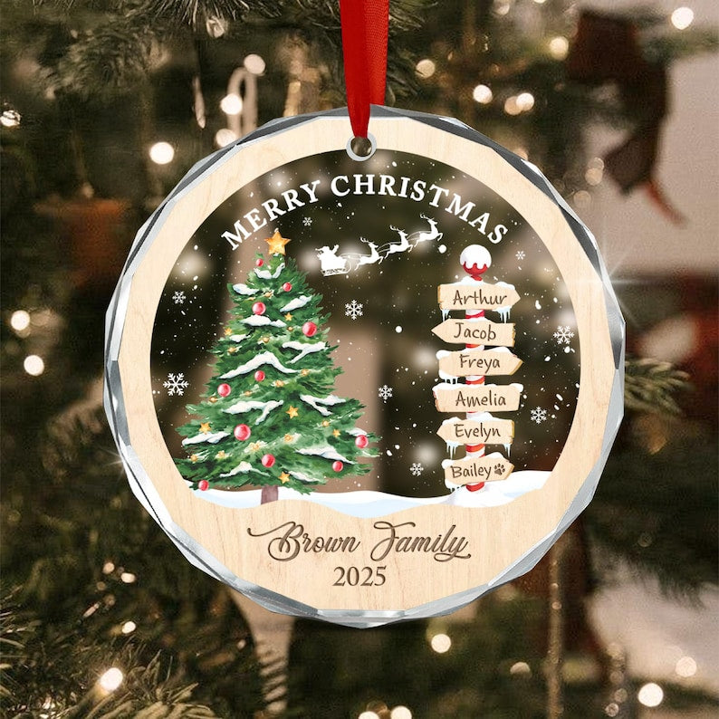 A round, faceted glass Christmas ornament with a red ribbon hanger. The design features a decorated pine tree, a silhouette of Santa in a sleigh, and a signpost with the names Arthur, Jacob, Freya, Amelia, Evelyn, and Bailey.