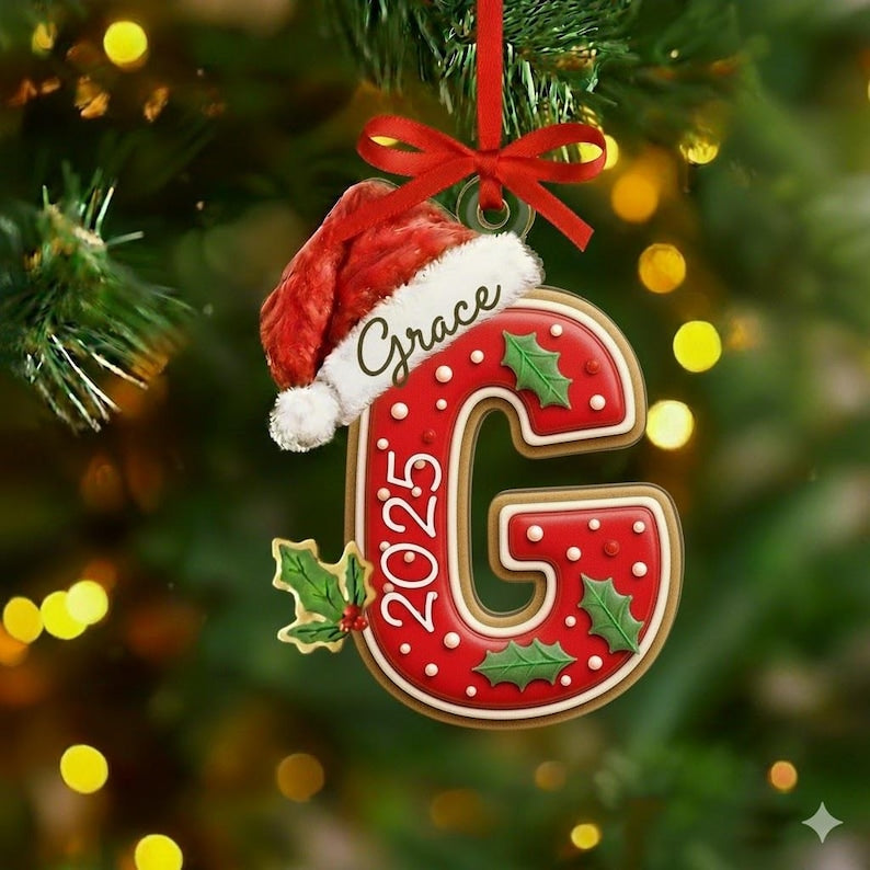 A red and white letter G Christmas ornament featuring a Santa hat on top, green holly leaves, and the text Grace and 2025. The ornament is suspended by a red ribbon and has a glossy finish with white polka dots on the red surface.