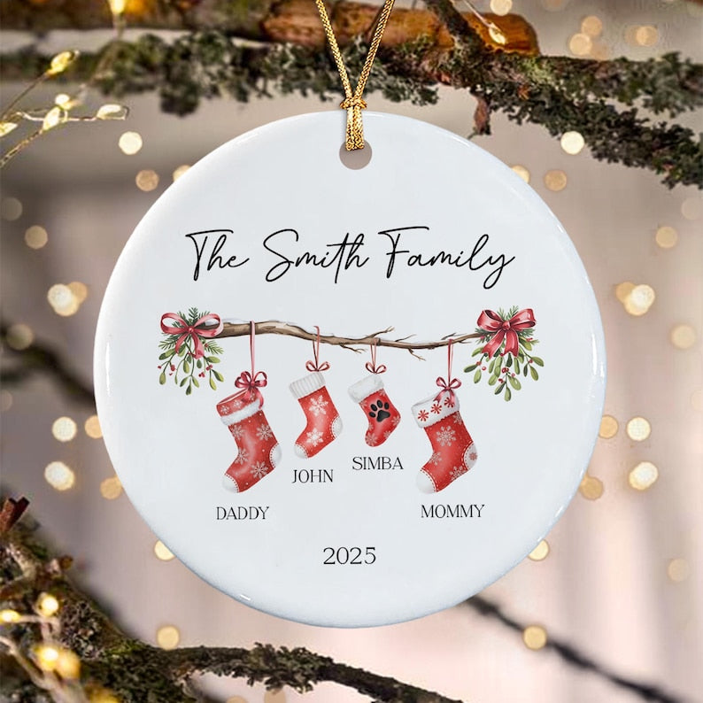 A round white ceramic Christmas ornament featuring a branch with four hanging red stockings. The text reads The Smith Family, Daddy, John, Simba, Mommy, and 2025. The stockings have snowflake patterns and a paw print on one.