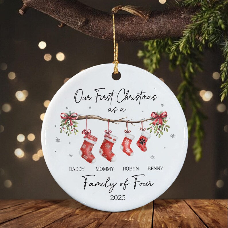 A round white ceramic Christmas ornament featuring the text Our First Christmas as a Family of Four 2025. The design includes a branch with four hanging red stockings, mistletoe, and the names Daddy, Mommy, Robyn, and Benny.