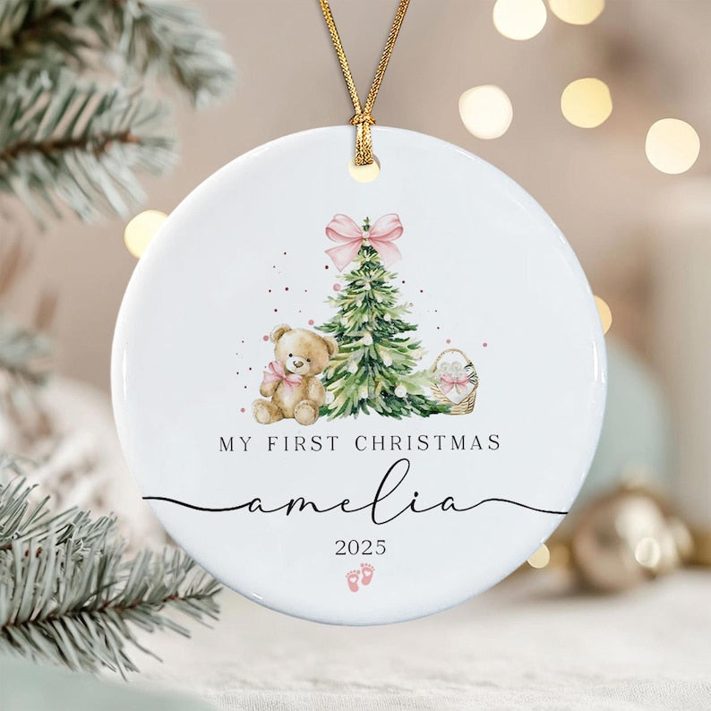 A round white ceramic ornament hanging from a gold string. The design features a watercolor teddy bear, a Christmas tree with a pink bow, and a basket.
