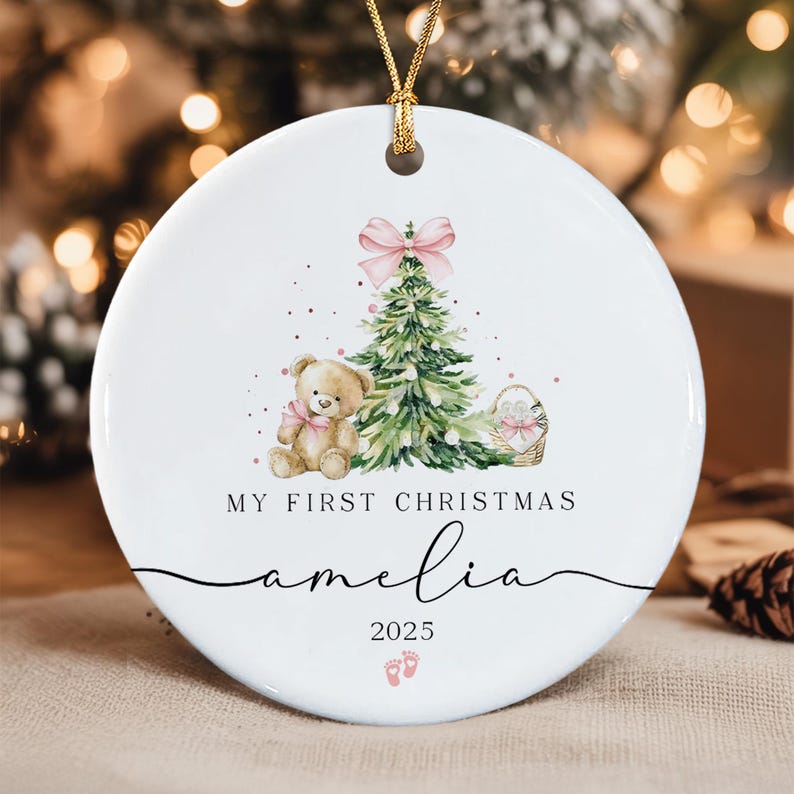 A round white ceramic ornament with a gold hanging cord. The design features a green Christmas tree with a pink bow, a teddy bear with a pink bow, and a basket.