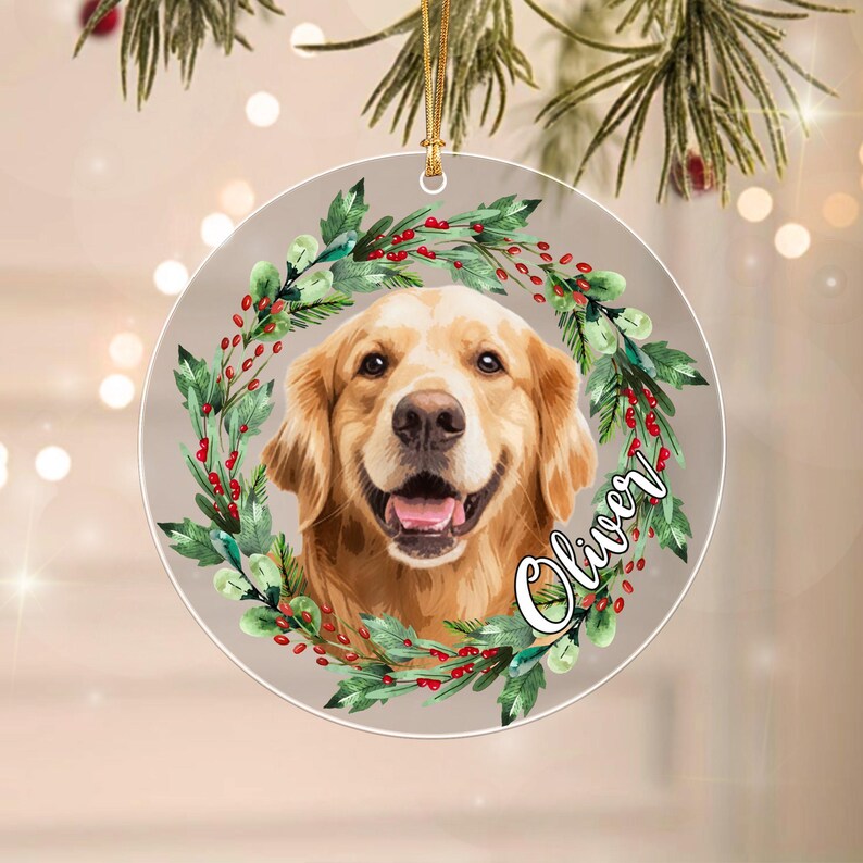 A round, flat ornament featuring a golden retriever portrait centered within a wreath of green leaves and red berries. The name Oliver is written in white cursive script on the right side. A gold string is attached to the top.