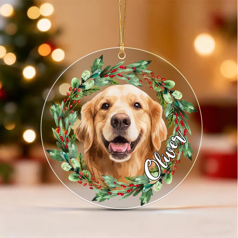 A circular clear acrylic ornament featuring a golden retriever portrait framed by a wreath of green leaves and red berries. The name Oliver is printed in white script on the right side.