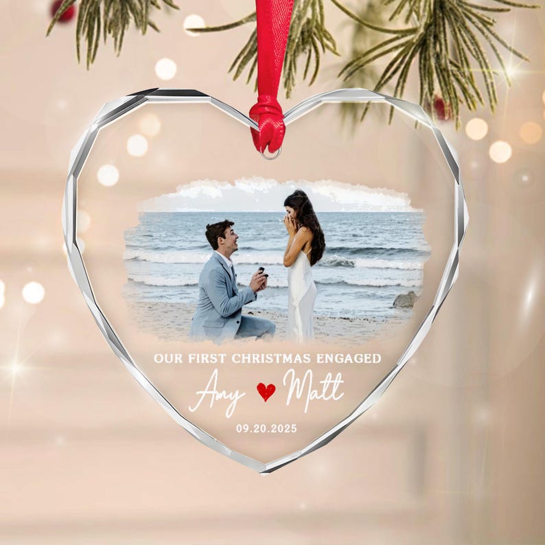 A heart-shaped clear glass ornament with faceted edges, featuring a printed photograph of a couple on a beach. The text reads OUR FIRST CHRISTMAS ENGAGED, Amy, Matt, and 09.20.2025, with a red heart icon.