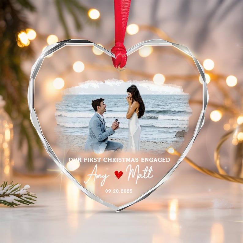 A heart-shaped clear acrylic ornament with faceted edges, featuring a printed photograph of a marriage proposal on a beach. The text reads OUR FIRST CHRISTMAS ENGAGED, Amy, a red heart icon, Matt, and the date 09.20.2025.