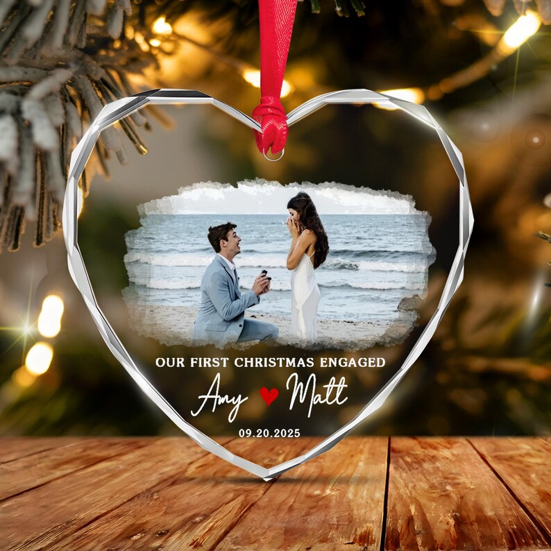 A heart-shaped clear glass ornament with faceted edges, featuring a printed photograph of a marriage proposal on a beach. The text reads OUR FIRST CHRISTMAS ENGAGED, Amy, a red heart icon, Matt, and the date 09.20.2025.