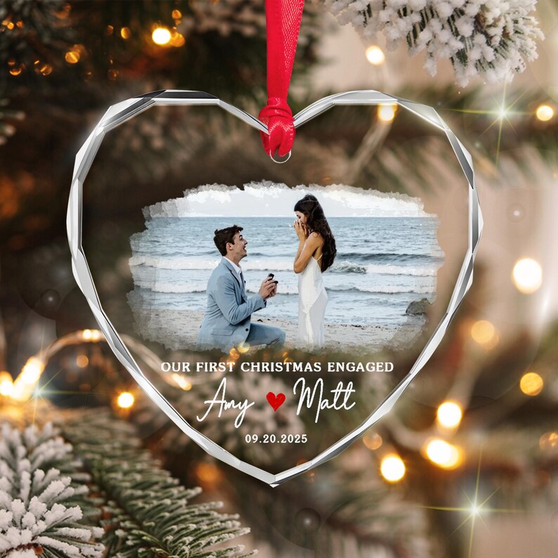 A heart-shaped clear glass ornament with faceted edges, featuring a printed photograph of a couple on a beach. The text reads: OUR FIRST CHRISTMAS ENGAGED, Amy, Matt, and 09.20.2025, with a small red heart icon.