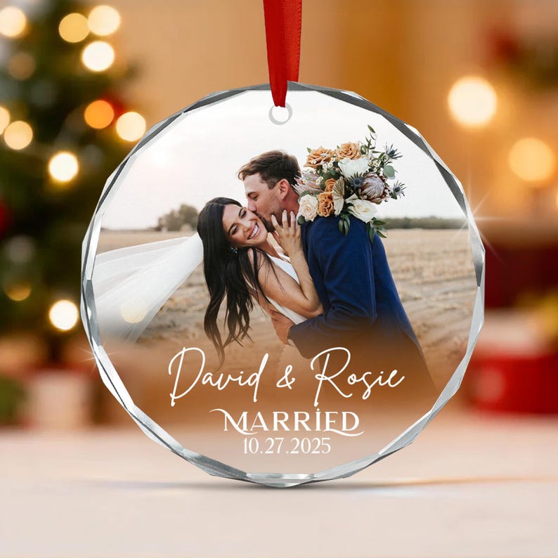 A circular faceted glass ornament featuring a printed photograph of a couple embracing outdoors. The text reads David & Rosie MARRIED 10.27.2025 in white script and serif fonts. A red ribbon is attached to the top for hanging.