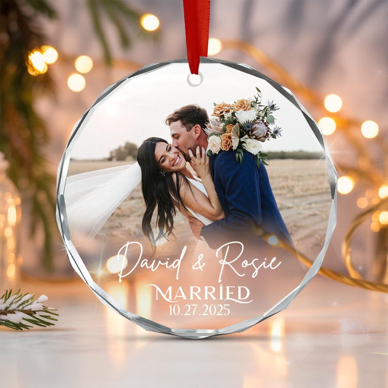 A circular faceted glass ornament featuring a printed photograph of a couple embracing with a bouquet of flowers. The text David & Rosie, MARRIED, and 10.27.2025 is printed in white script and serif fonts.