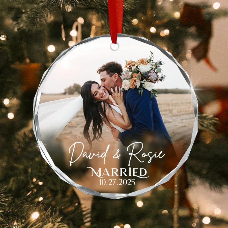 A round, faceted clear glass Christmas ornament featuring a printed photograph of a couple embracing outdoors. The text "David & Rosie" is printed in white script, followed by "MARRIED" and the date "10.27.2025" in white text.