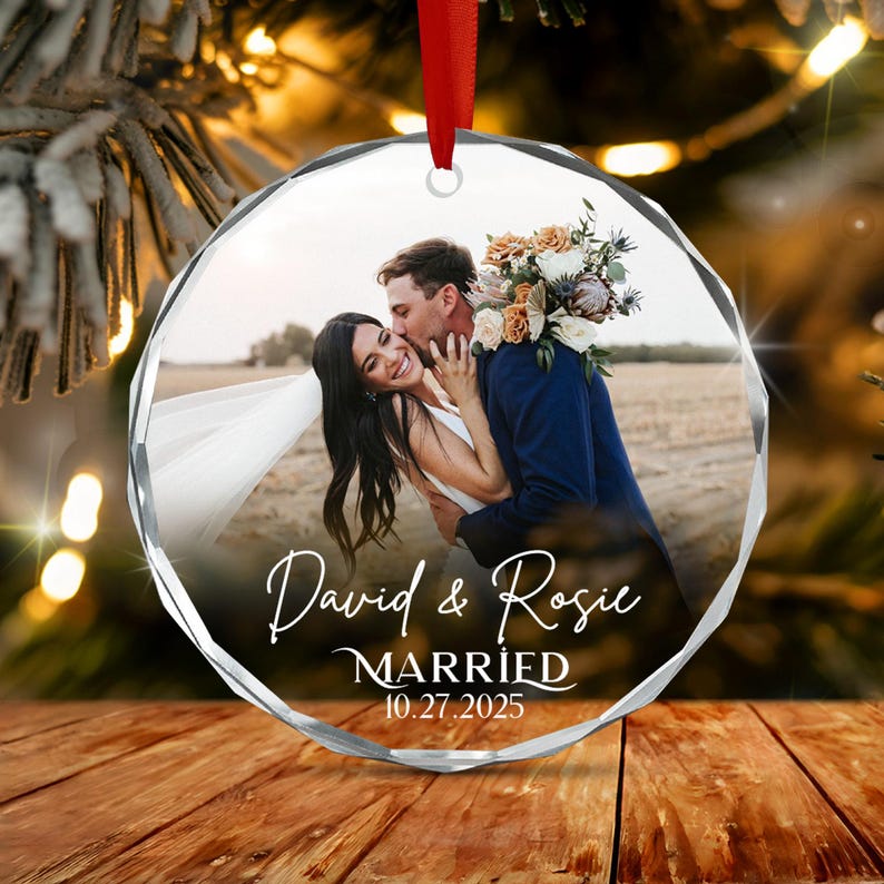 A circular faceted glass ornament featuring a photograph of a couple embracing, with the bride in a white dress and the groom in a navy suit. The text "David & Rosie MARRIED 10.27.2025" is printed in white script and serif font.