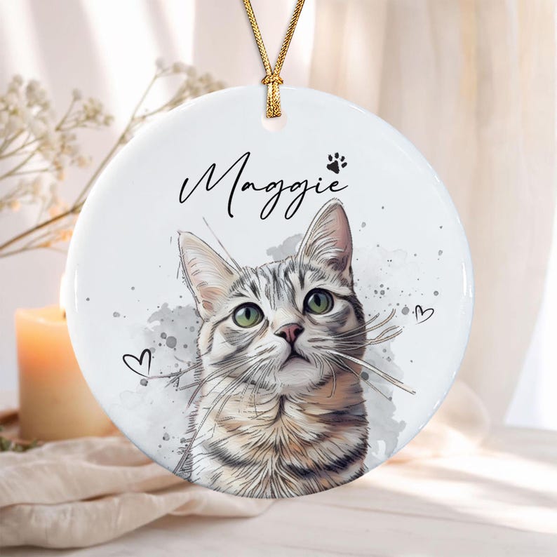 A round white ceramic ornament featuring a watercolor-style illustration of a tabby cat with green eyes. The name Maggie is printed in black script above the cat, accompanied by a small paw print and two small heart outlines.