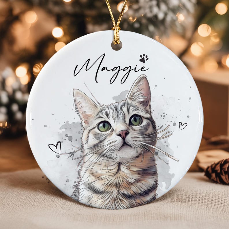 A round white ceramic ornament featuring a watercolor-style illustration of a grey tabby cat with green eyes. The name Maggie is printed in black script above the cat, accompanied by a small paw print and two hand-drawn hearts.
