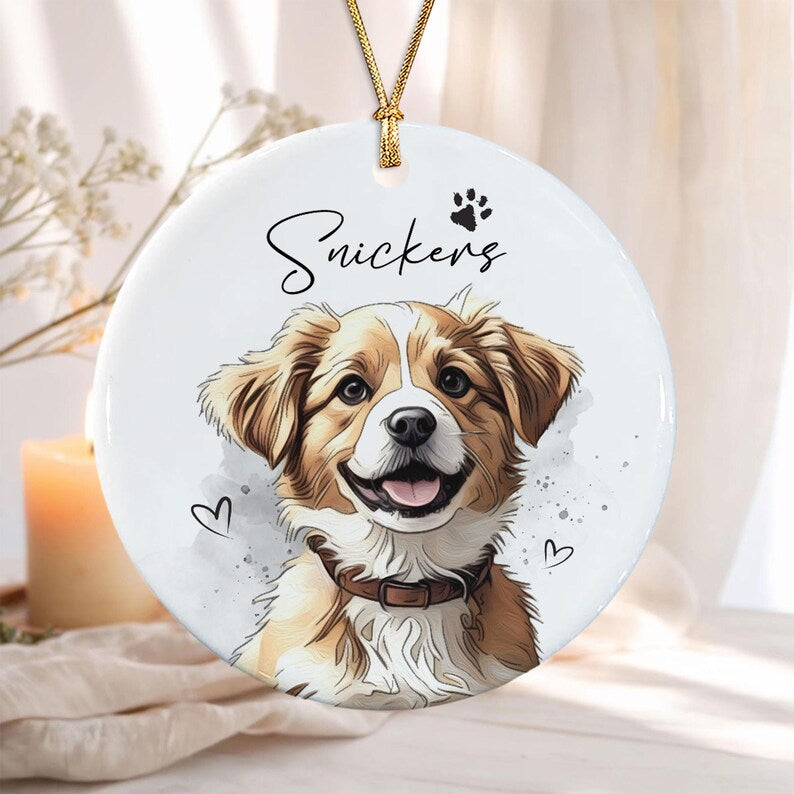 A round white ceramic ornament featuring a colorful illustration of a brown and white dog with a collar. The name Snickers is printed in black script above the dog, accompanied by a small paw print and two small heart outlines.