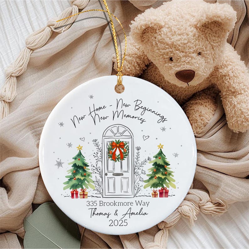 A round white ceramic ornament with a gold string featuring a printed illustration of a front door with a wreath, two Christmas trees, and the text New Home - New Beginnings New Memories, 335 Brookmore Way, Thomas & Amelia, 2025.