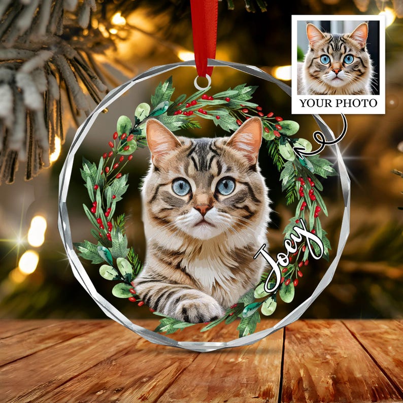 A circular, faceted glass Christmas ornament featuring a central portrait of a brown tabby cat with blue eyes, framed by a green holly wreath with red berries.