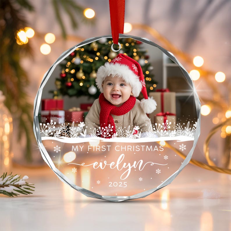 A round, faceted glass Christmas ornament featuring a photo of a baby wearing a red Santa hat and scarf. The ornament includes white printed text that reads MY FIRST CHRISTMAS, the name evelyn, and the year 2025 with snowflakes.