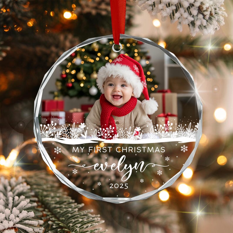A circular glass Christmas ornament featuring a photo of a baby wearing a red Santa hat and scarf. The ornament includes white printed text that reads MY FIRST CHRISTMAS, the name evelyn, and the year 2025 with snowflake accents.