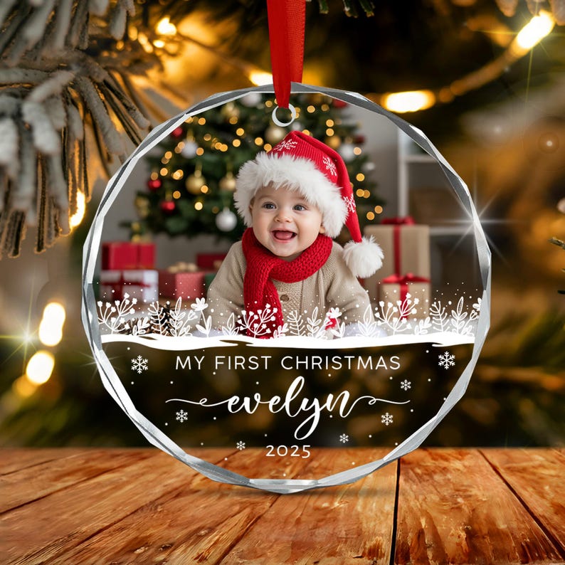 A circular glass Christmas ornament featuring a photo of a baby wearing a red Santa hat and scarf. The ornament includes white printed text that reads MY FIRST CHRISTMAS, the name evelyn, and the year 2025 with snowflake accents.