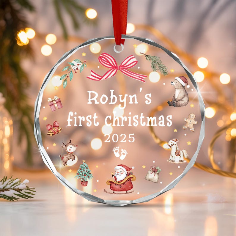 A round, clear acrylic Christmas ornament with a red ribbon hanger. The text reads "Robyn's first christmas 2025" with illustrations of a bear, gingerbread man, rocking horse, Santa in a sleigh, a tree, and baby footprints.