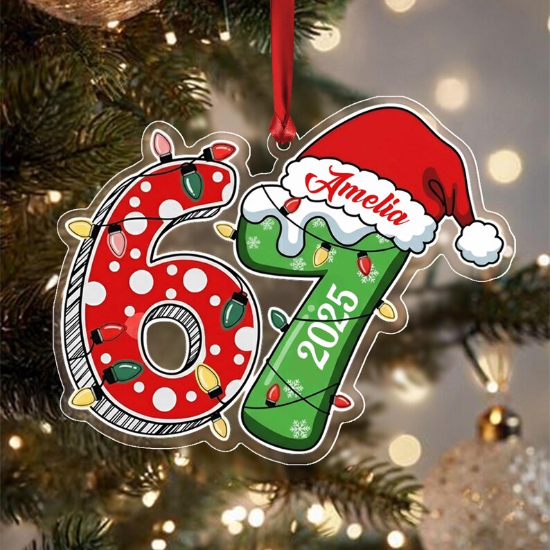 A custom Christmas ornament shaped like the number 67. The number 6 is red with white polka dots, and the number 7 is green with white snowflakes. A red Santa hat sits atop the 7, featuring the name Amelia and the year 2025.