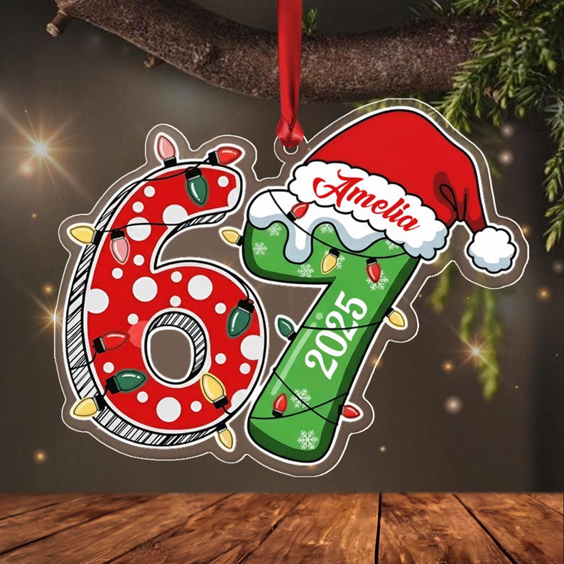 A flat hanging ornament shaped like the number 67. The 6 is red with white polka dots, and the 7 is green with white snowflakes. A red Santa hat sits atop the 7, which features the name Amelia and the year 2025 in white text.