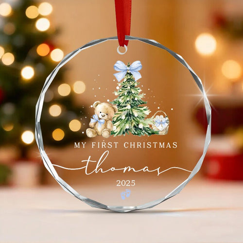 A circular clear glass ornament with faceted edges, featuring a printed illustration of a teddy bear, a Christmas tree, and a basket. The text reads MY FIRST CHRISTMAS, thomas, and 2025, with two small blue baby footprints.