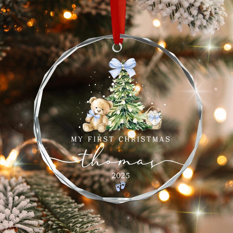 A circular clear glass Christmas ornament with a red ribbon hanger. The design features a watercolor teddy bear, a green Christmas tree, a basket, and a light blue bow.