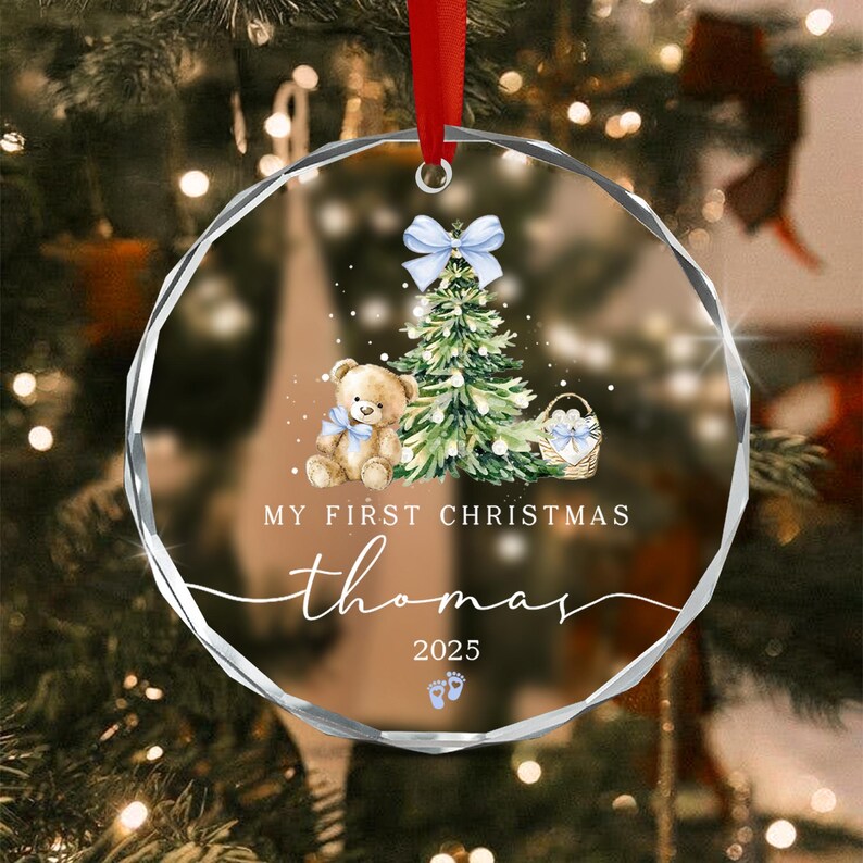 A round, clear acrylic Christmas ornament featuring a printed illustration of a teddy bear, a decorated pine tree, and a basket. The text reads MY FIRST CHRISTMAS, thomas, and 2025, with two small blue baby footprints below.