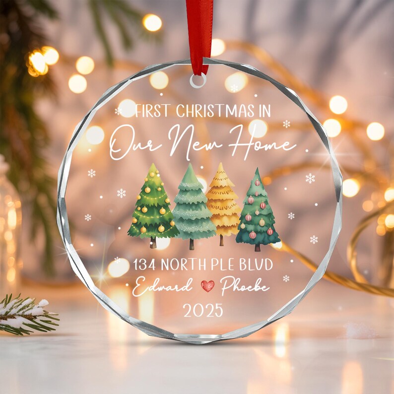 A circular clear glass Christmas ornament with a red ribbon hanger. It features the text FIRST CHRISTMAS IN Our New Home, 134 NORTH PLE BLVD, Edward, a red heart, Phoebe, and 2025.