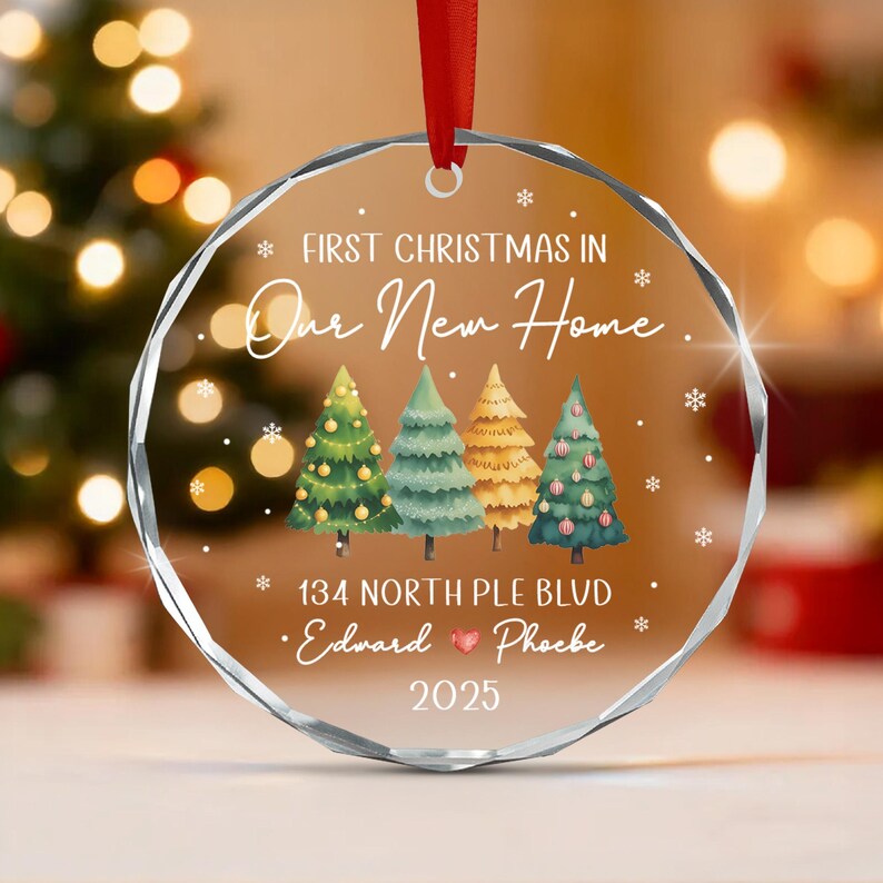A circular clear glass ornament with a faceted edge, featuring a red ribbon hanger. The text reads FIRST CHRISTMAS IN Our New Home, 134 NORTH PLE BLVD, Edward and Phoebe, and 2025. Four illustrated Christmas trees are centered.
