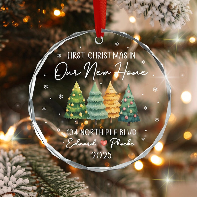 A round, clear, faceted glass ornament hanging from a red ribbon. It features the text "FIRST CHRISTMAS IN Our New Home", "134 NORTH PLE BLVD", "Edward ❤ Phoebe", and "2025" with four illustrated Christmas trees and snowflakes.