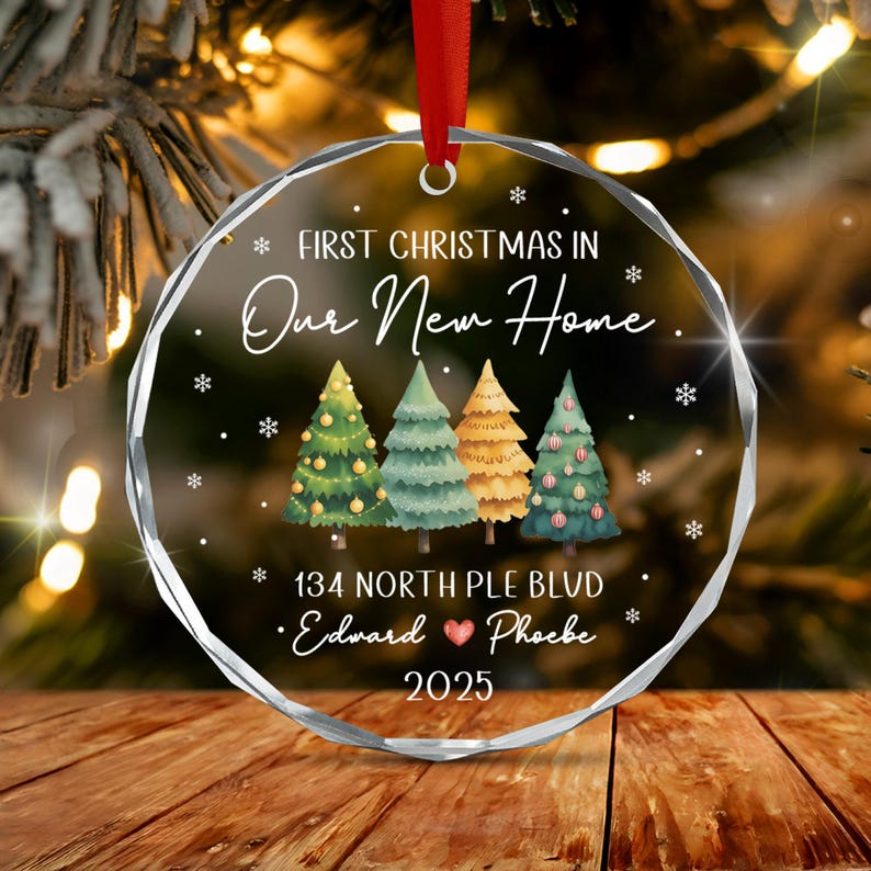 A round, clear acrylic Christmas ornament with a faceted edge, featuring the text "FIRST CHRISTMAS IN Our New Home", "134 NORTH PLE BLVD", "Edward ❤ Phoebe", and "2025".