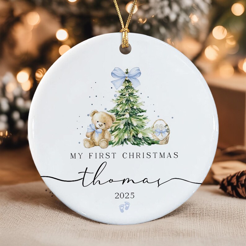 A round white ceramic ornament featuring a watercolor illustration of a teddy bear, a Christmas tree with a light blue bow, and a basket.