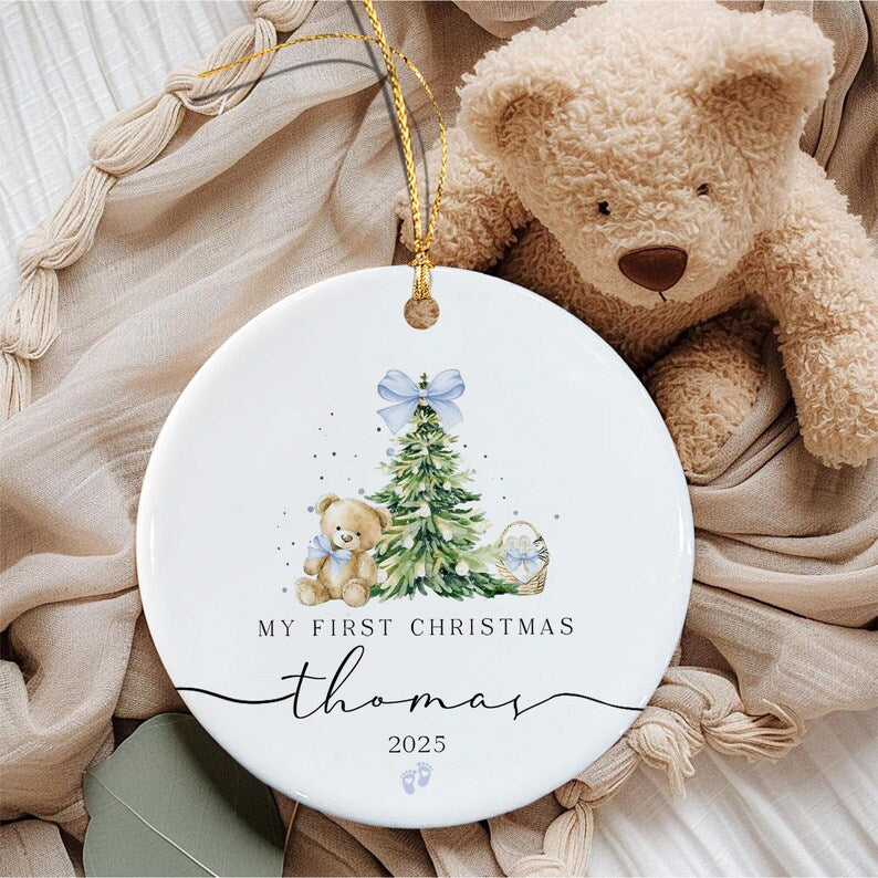 A round white ceramic ornament featuring a watercolor illustration of a green Christmas tree, a light brown teddy bear with a blue bow, and a small basket.