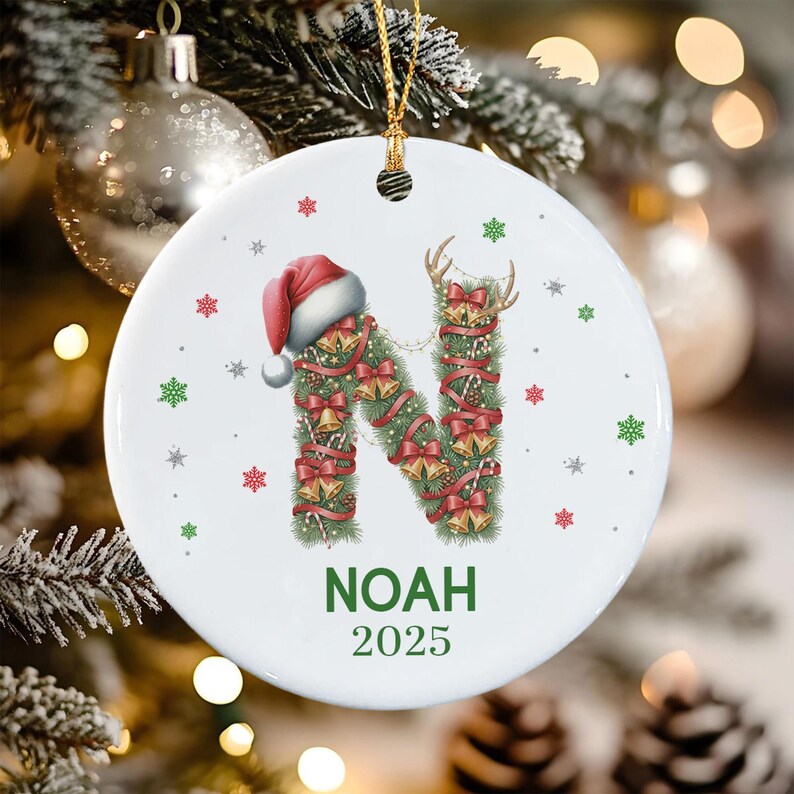A round white ceramic ornament featuring a large letter N decorated with green pine needles, red ribbons, gold bells, and a Santa hat.