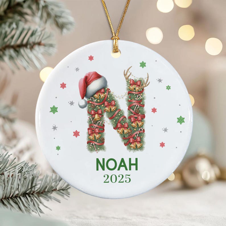 A round white ceramic ornament featuring a large letter N shaped like a pine branch, decorated with red ribbons, gold bells, and a Santa hat.