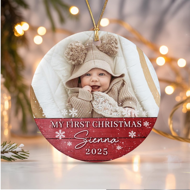 A round ceramic Christmas ornament featuring a photograph of a baby wearing a beige hooded sweater with pom-poms. The bottom section has a red wood-grain background with white text reading MY FIRST CHRISTMAS, Sienna, and 2025.