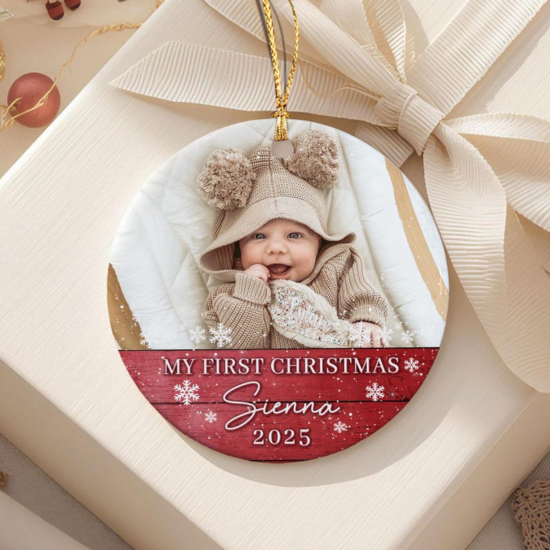A round ceramic Christmas ornament featuring a photograph of a baby wearing a beige hooded sweater with pom-poms.